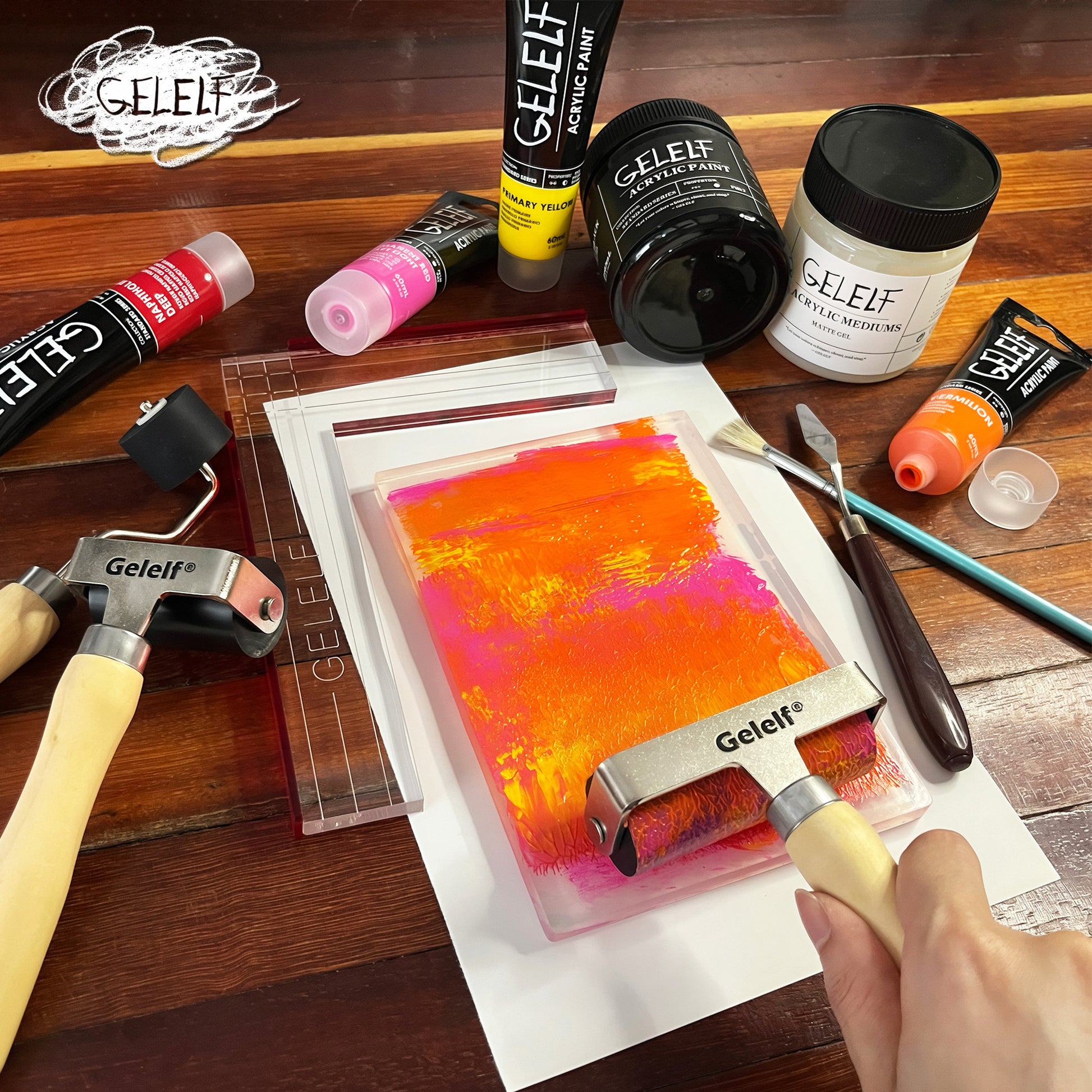 Image of an gelli print artist spreading gelelf printmaking paint onto a gelelf 5 x 7 inch gel plate, with gelelf gel medium, acrylic paint, gelelf brayers, and the gelelf perfect positioning ruler placed around it.